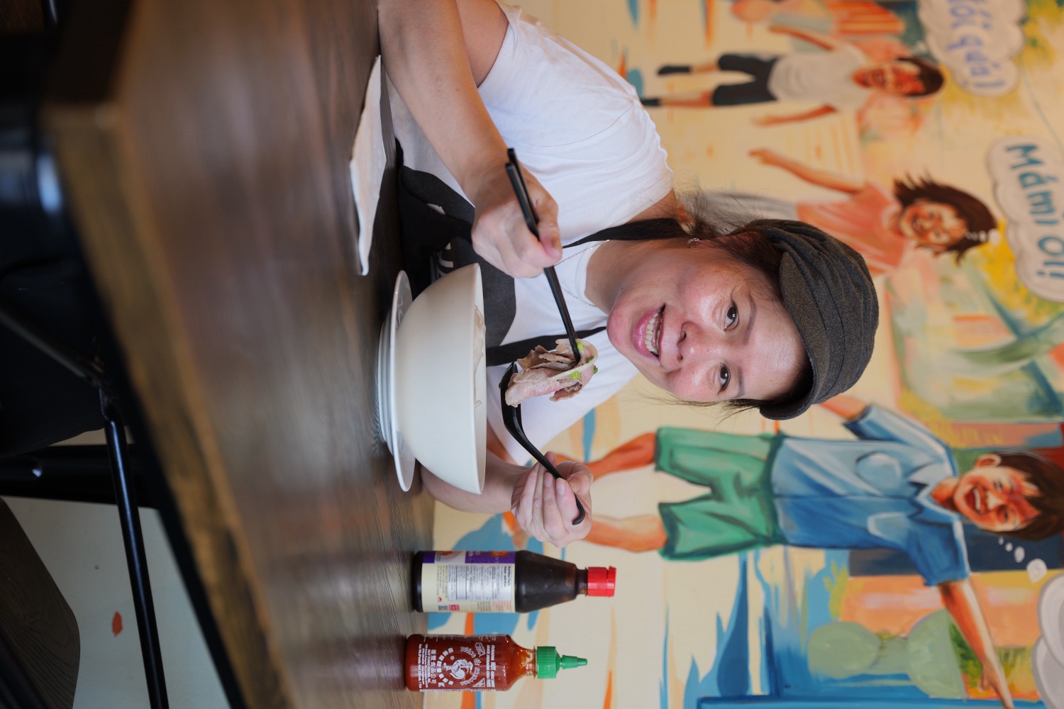 Mámì enjoying a bowl of pho at the table, in front of the Chào Mámì mural